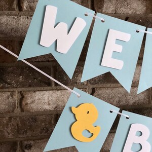 Rubber duck Welcome Baby sign. The flags are made with light blue cardstock backgrounds and white letters. The letters are attached with 3d tape to give it dimension. The two end flags are decorated with yellow rubber ducks. These are strung on a coordinating satin ribbon with plenty of extra to hang the banner.