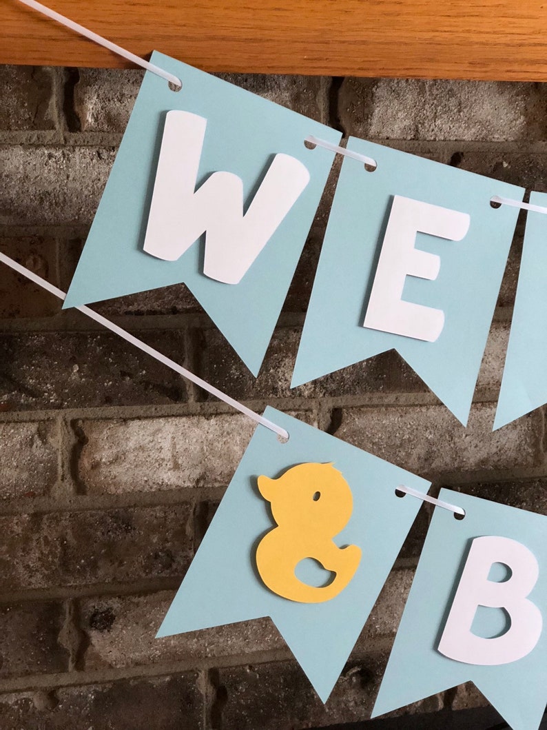 Rubber duck Welcome Baby sign. The flags are made with light blue cardstock backgrounds and white letters. The letters are attached with 3d tape to give it dimension. The two end flags are decorated with yellow rubber ducks. These are strung on a coordinating satin ribbon with plenty of extra to hang the banner.