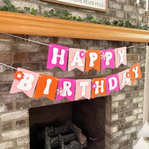 May include: A two-row banner spelling HAPPY BIRTHDAY with white letters on alternating pink, light pink, and orange pennants. Each pennant features a small flower cutout in a contrasting color, strung together on a thin white cord.