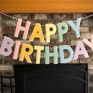 HAPPY BIRTHDAY banner made with pastel cardstock. Background made from white cardstock. Letters are cut out of different colors of sturdy cardstock. These are attached with 3d tape. Letters are strung together with white ribbon. There is extra ribbon on the ends for you to hang it up.