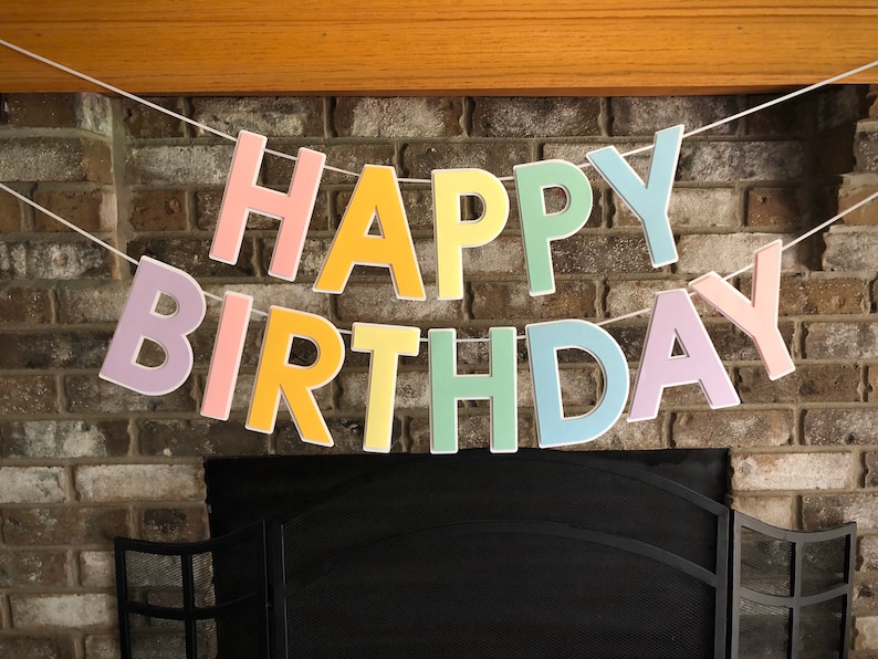 HAPPY BIRTHDAY banner made with pastel cardstock. Background made from white cardstock. Letters are cut out of different colors of sturdy cardstock. These are attached with 3d tape. Letters are strung together with white ribbon. There is extra ribbon on the ends for you to hang it up.