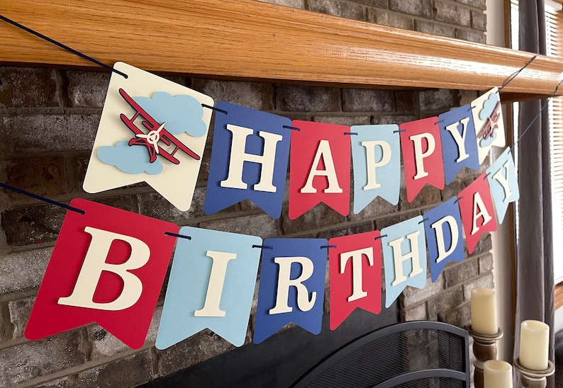 Vintage airplane Happy Birthday banner. Alternating navy blue, red and light blue cardstock flags. Ivory letters are attached with 3D tape. 3D airplanes decorate the two end flags. The banner is strung on a matching ribbon ready to hang.