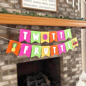 May include: A two-tiered banner featuring the text TWOTTI FRUTTI in white block letters on colorful pennants. The banner includes cutouts of a pineapple, a dragon fruit slice, and cherries, alternating between pink, green, orange, and yellow colors.