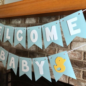 Rubber duck Welcome Baby sign. The flags are made with light blue cardstock backgrounds and white letters. The letters are attached with 3d tape to give it dimension. The two end flags are decorated with yellow rubber ducks. These are strung on a coordinating satin ribbon with plenty of extra to hang the banner.