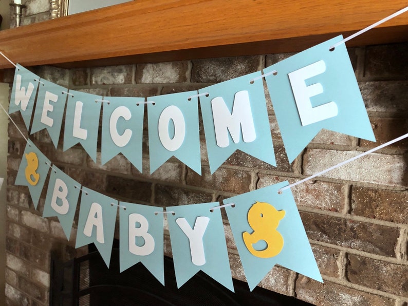Rubber duck Welcome Baby sign. The flags are made with light blue cardstock backgrounds and white letters. The letters are attached with 3d tape to give it dimension. The two end flags are decorated with yellow rubber ducks. These are strung on a coordinating satin ribbon with plenty of extra to hang the banner.