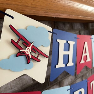 Vintage airplane Happy Birthday banner. Alternating navy blue, red and light blue cardstock flags. Ivory letters are attached with 3D tape. 3D airplanes decorate the two end flags. The banner is strung on a matching ribbon ready to hang.