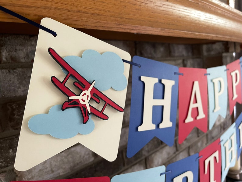 Vintage airplane Happy Birthday banner. Alternating navy blue, red and light blue cardstock flags. Ivory letters are attached with 3D tape. 3D airplanes decorate the two end flags. The banner is strung on a matching ribbon ready to hang.