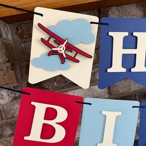 Vintage airplane Happy Birthday banner. Alternating navy blue, red and light blue cardstock flags. Ivory letters are attached with 3D tape. 3D airplanes decorate the two end flags. The banner is strung on a matching ribbon ready to hang.