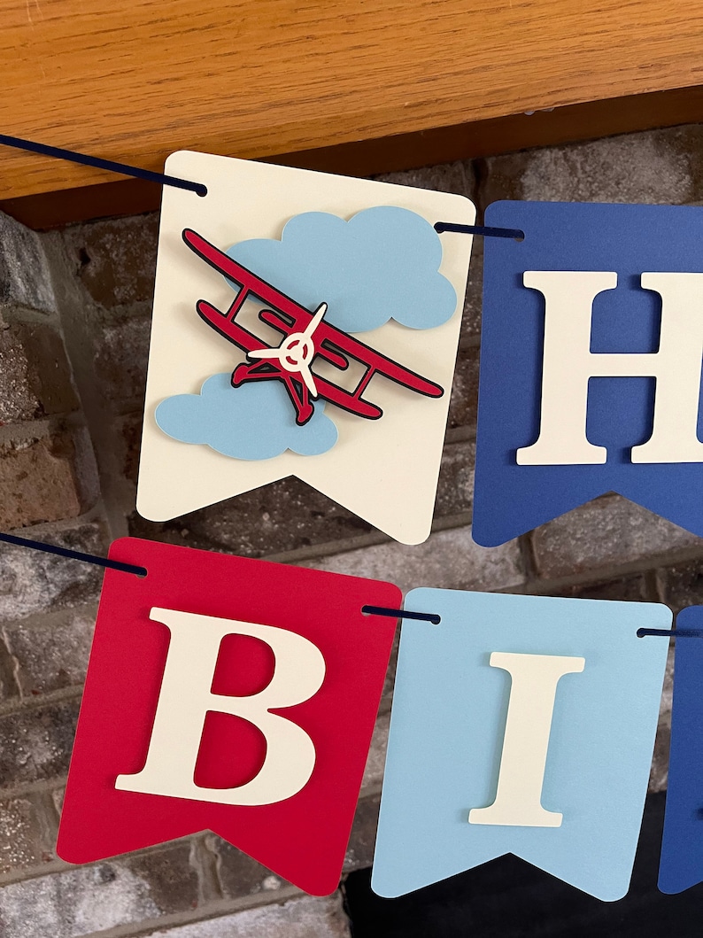 Vintage airplane Happy Birthday banner. Alternating navy blue, red and light blue cardstock flags. Ivory letters are attached with 3D tape. 3D airplanes decorate the two end flags. The banner is strung on a matching ribbon ready to hang.
