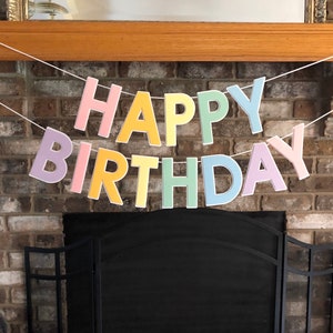 HAPPY BIRTHDAY banner made with pastel cardstock. Background made from white cardstock. Letters are cut out of different colors of sturdy cardstock. These are attached with 3d tape. Letters are strung together with white ribbon. There is extra ribbon on the ends for you to hang it up.