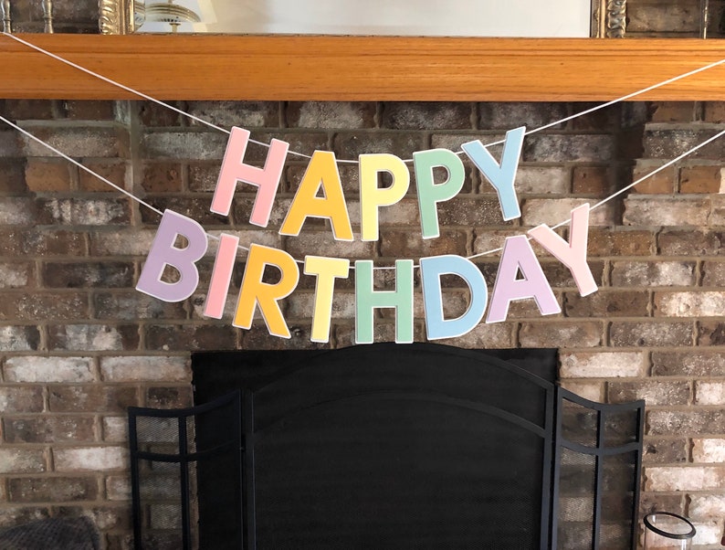 HAPPY BIRTHDAY banner made with pastel cardstock. Background made from white cardstock. Letters are cut out of different colors of sturdy cardstock. These are attached with 3d tape. Letters are strung together with white ribbon. There is extra ribbon on the ends for you to hang it up.