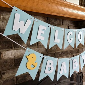 Rubber duck Welcome Baby sign. The flags are made with light blue cardstock backgrounds and white letters. The letters are attached with 3d tape to give it dimension. The two end flags are decorated with yellow rubber ducks. These are strung on a coordinating satin ribbon with plenty of extra to hang the banner.
