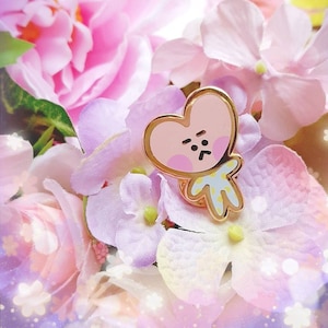 May include: A gold-colored pin featuring a heart-shaped character with a light pink face and a grumpy expression. The character has a small body in a light blue outfit with yellow spots. The pin is set against a backdrop of pink and purple flowers.