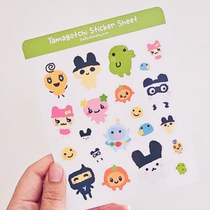 Puede incluir: Una hoja de pegatinas de Tamagotchi con varios personajes de la popular franquicia de mascotas virtuales. Las pegatinas están impresas en papel blanco con un borde verde y el texto "Tamagotchi Sticker Sheet" y "lilcakecake.etsy.com".