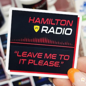May include: A black and red sticker with the text "Hamilton Radio" and a yellow shield with the letter "A" inside. The sticker also has a red equalizer graphic and the text "Leave me to it please."
