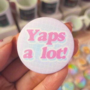 Op de afbeelding: Een ronde, witte button met de tekst "Yaps a lot!" in roze, omlijnd met een lichter blauw. De button heeft een subtiel rasterpatroon en wordt in een hand gehouden, met andere buttons op de achtergrond.