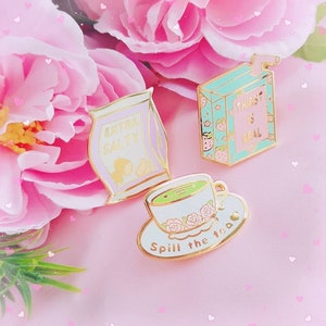 May include: Three enamel pins with a pastel colour scheme. The first pin is a cartoon image of a bag of crisps with the text "Extra Salty". The second pin is a cartoon image of a cup of tea with the text "Spill the tea". The third pin is a cartoon image of a juice carton with the text "Thirst is Real".