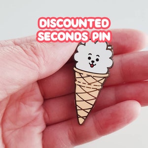Op de afbeelding: Speld in de vorm van een ijshoorntje met een wit, pluizig gezicht en een lachende uitdrukking. De kegel is bruin met bruine lijnen. De tekst "DISCOUNTED SECONDS PIN" staat in een rode tekstballon.