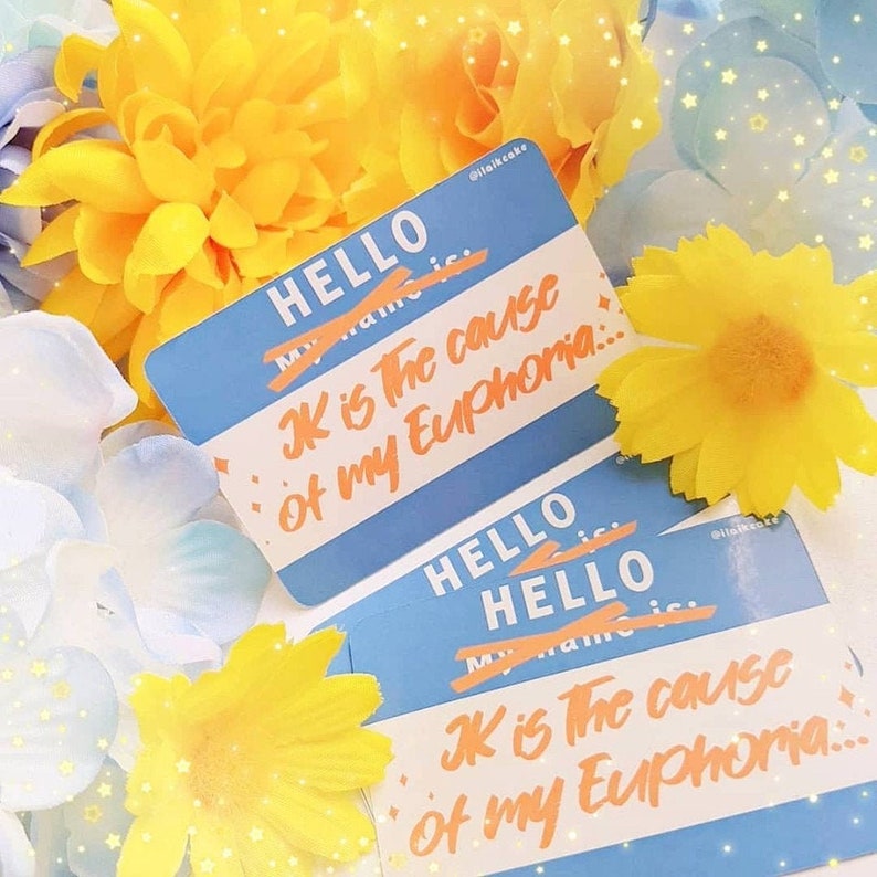 May include: Blue name tags with the word "HELLO" at the top and the text "JK is the cause of my Euphoria..." in orange. The background includes yellow and white flowers.