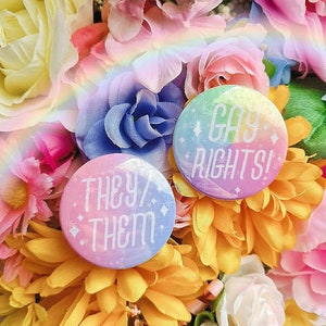 May include: Two round buttons with rainbow-colored backgrounds and white text. The first button says "They/Them" and the second button says "Gay Rights!"
