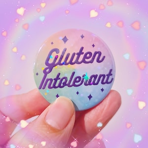 Może przedstawiać: Okrągła, pastelowa przypinka z napisem "Gluten Intolerant" w fioletowym skrypcie. Przypinka ma holograficzne, błyszczące wykończenie z akcentami gwiazd i diamentów. Tło to miękka, marzycielska mieszanka kolorów.