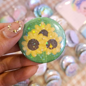 Peut inclure: Un bouton vert avec un motif de tournesol jaune. Le tournesol a un effet brillant et est entouré de petites étoiles blanches.