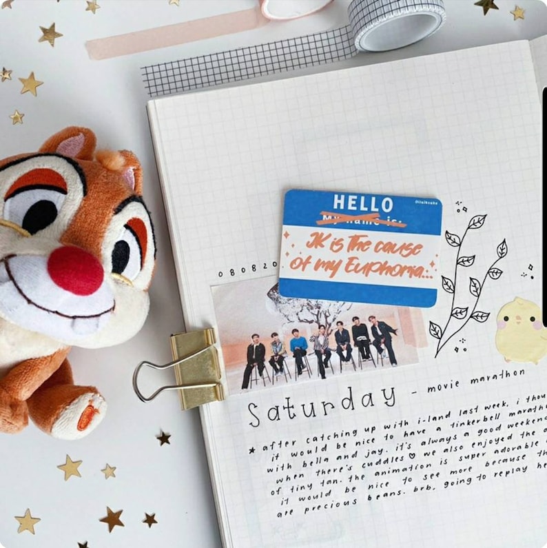 May include: A scrapbook page with a plush Chipmunk toy, a blue name tag that says "JK is the cause of my Euphoria", a photo of a group of people, and the word "Saturday". Decorative stars and tape are also visible.