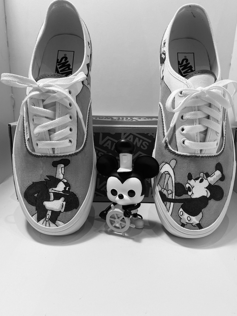 May include: Pair of gray and white Vans sneakers with custom Mickey Mouse artwork. One shoe features Pete, the other Mickey Mouse at the helm of a ship. A Mickey Mouse figurine is in front of the shoes.