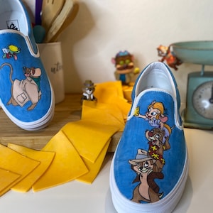 Peut inclure: Paire de baskets bleues à enfiler avec des illustrations de personnages de dessins animés. Une chaussure représente un personnage de souris, l'autre des personnages de Tic et Tac. Semelles blanches et une planche à découper en bois sont également visibles.