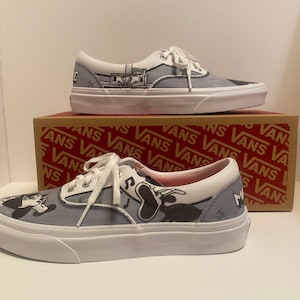 May include: Pair of gray and white Vans sneakers with custom black and white cartoon artwork. The shoes have white laces and soles. The Vans box is visible in the background.