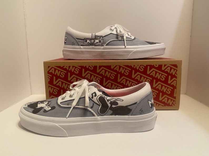 May include: Pair of gray and white Vans sneakers with custom black and white cartoon artwork. The shoes have white laces and soles. The Vans box is visible in the background.