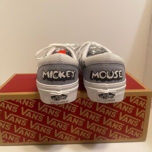 May include: Pair of white and gray Vans sneakers with "MICKEY" and "MOUSE" printed on the heels. The shoes are on top of a brown and red Vans box. The shoes have white laces and a white sole.