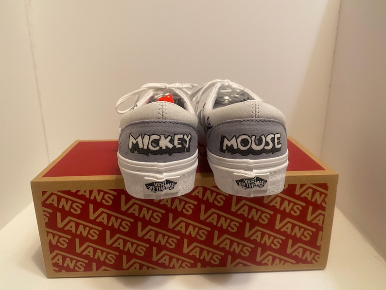 May include: Pair of white and gray Vans sneakers with "MICKEY" and "MOUSE" printed on the heels. The shoes are on top of a brown and red Vans box. The shoes have white laces and a white sole.