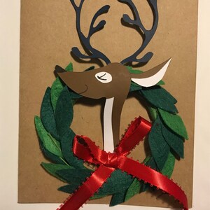 May include: A handmade Christmas card featuring a brown reindeer head with navy blue antlers, a green felt wreath, and a red ribbon. The card is on a brown background.