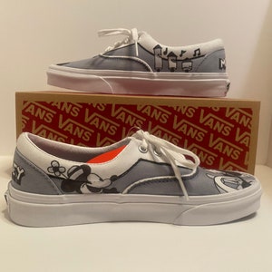 May include: Pair of gray Vans Era sneakers with white soles and laces. The shoes feature black and white cartoon graphics and the word "Mickey". The shoes are displayed on top of a Vans box.