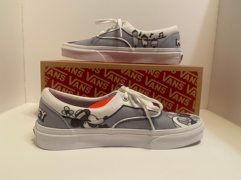 May include: Pair of gray Vans Era sneakers with white soles and laces. The shoes feature black and white cartoon graphics and the word "Mickey". The shoes are displayed on top of a Vans box.