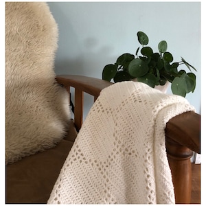 Può includere: Una coperta bianca all'uncinetto drappeggiata su una sedia di legno con un cuscino soffice in pelliccia beige. Una pianta in vaso con foglie verdi rotonde si trova sul bracciolo della sedia.