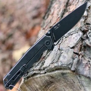 May include: A black folding knife with a matte finish. The blade is angled and features the text "P801-SB". The handle has a clip for secure carrying. The knife is shown against a natural wood background.
