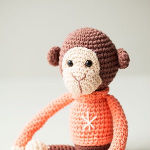 May include: A crocheted monkey toy wearing a coral sweater with a white snowflake design. The monkey has brown fur, a cream-colored face, and black eyes.