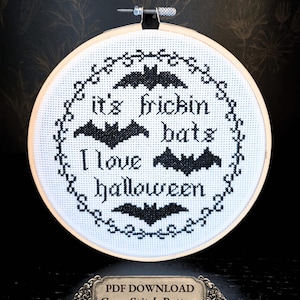 May include: A circular wooden embroidery hoop containing white fabric with a black cross-stitch design featuring four bats and the text "it's frickin bats I love halloween" enclosed in a decorative border. A label below reads "PDF DOWNLOAD Cross Stitch Pattern 6" Finished Size".