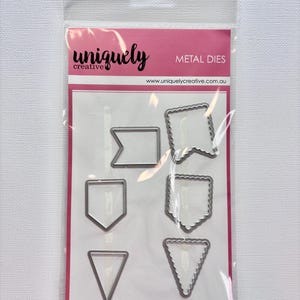 May include: A package of Uniquely Creative metal dies in various banner shapes. The package includes a rectangular, flag, pennant, and triangle die cuts. The packaging is pink and white with the brand name and website address printed on it.