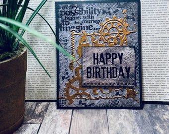 May include: A rectangular greeting card featuring a dark, textured background with gold-colored gear embellishments. The text reads, "Every possibility begins with the courage to imagine..." and "HAPPY BIRTHDAY" in bold, black, capitalized letters.