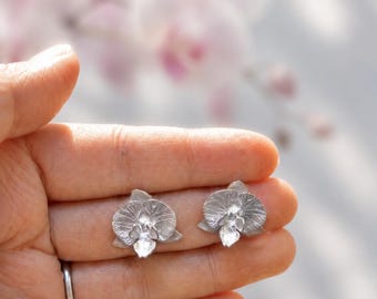 Może przedstawiać: Para srebrzystych kolczyków w kształcie kwiatów orchidei. Każdy kolczyk ma teksturowany wzór przypominający płatki z małym, przezroczystym, fasetowanym kamieniem osadzonym pośrodku, naśladującym wygląd kwitnącego kwiatu orchidei.