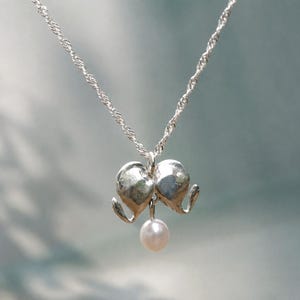 Op de afbeelding: Een zilveren ketting met een fijne gedraaide schakelketting en een hanger in de vorm van twee gestileerde metalen olifantenkoppen die naar elkaar toe kijken, met een kleine witte parel hangend in het midden tussen de twee slurfjes.