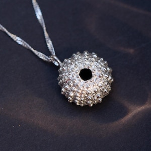 Puede incluir: Collar con colgante plateado. El colgante es un dije esférico texturizado con un agujero central, cubierto de pequeñas protuberancias. Cuelga de una delicada cadena trenzada. El collar se muestra sobre un fondo oscuro.