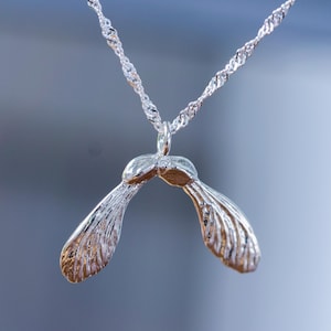 Puede incluir: Un collar de plata con un colgante con forma de semilla de arce. El colgante es detallado y tiene un diseño delicado e intrincado.