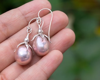 May include: A pair of silver earrings featuring teardrop-shaped, light pink pearls. The earrings have a floral design at the top and a hook closure. The pearls have a subtle luster, and the earrings are displayed against a blurred green background.