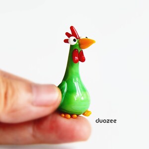 Peut inclure: Une figurine de coq en verre vert avec une cr&ecirc;te et un bec rouges, et de grands yeux blancs. Le coq est debout sur une surface blanche.