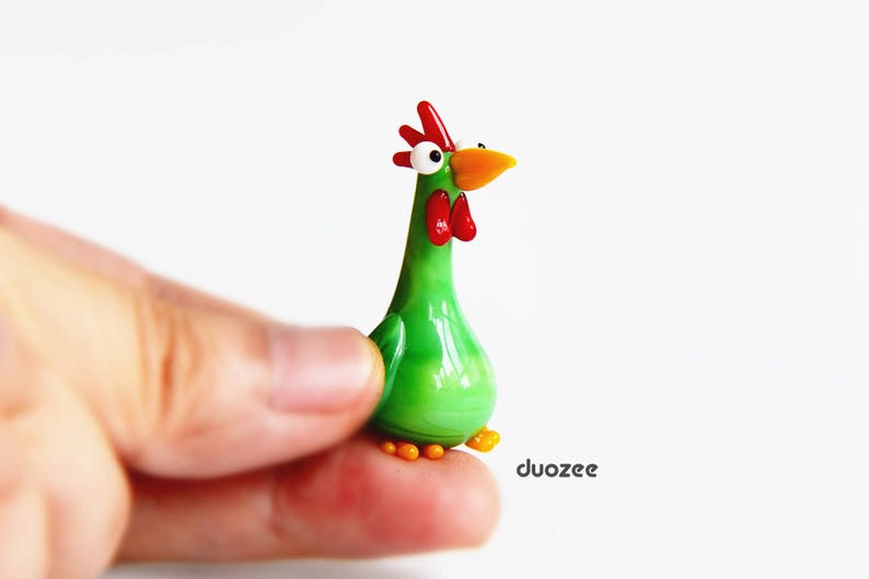 Peut inclure: Une figurine de coq en verre vert avec une cr&ecirc;te et un bec rouges, et de grands yeux blancs. Le coq est debout sur une surface blanche.