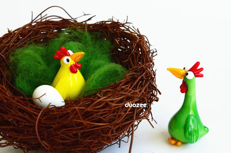 Peut inclure: Une figurine de poulet en verre jaune et rouge est plac&eacute;e dans un nid de brindilles brun avec de la mousse verte. Une figurine de poulet en verre vert avec des accents rouges se tient &agrave; l'ext&eacute;rieur du nid.